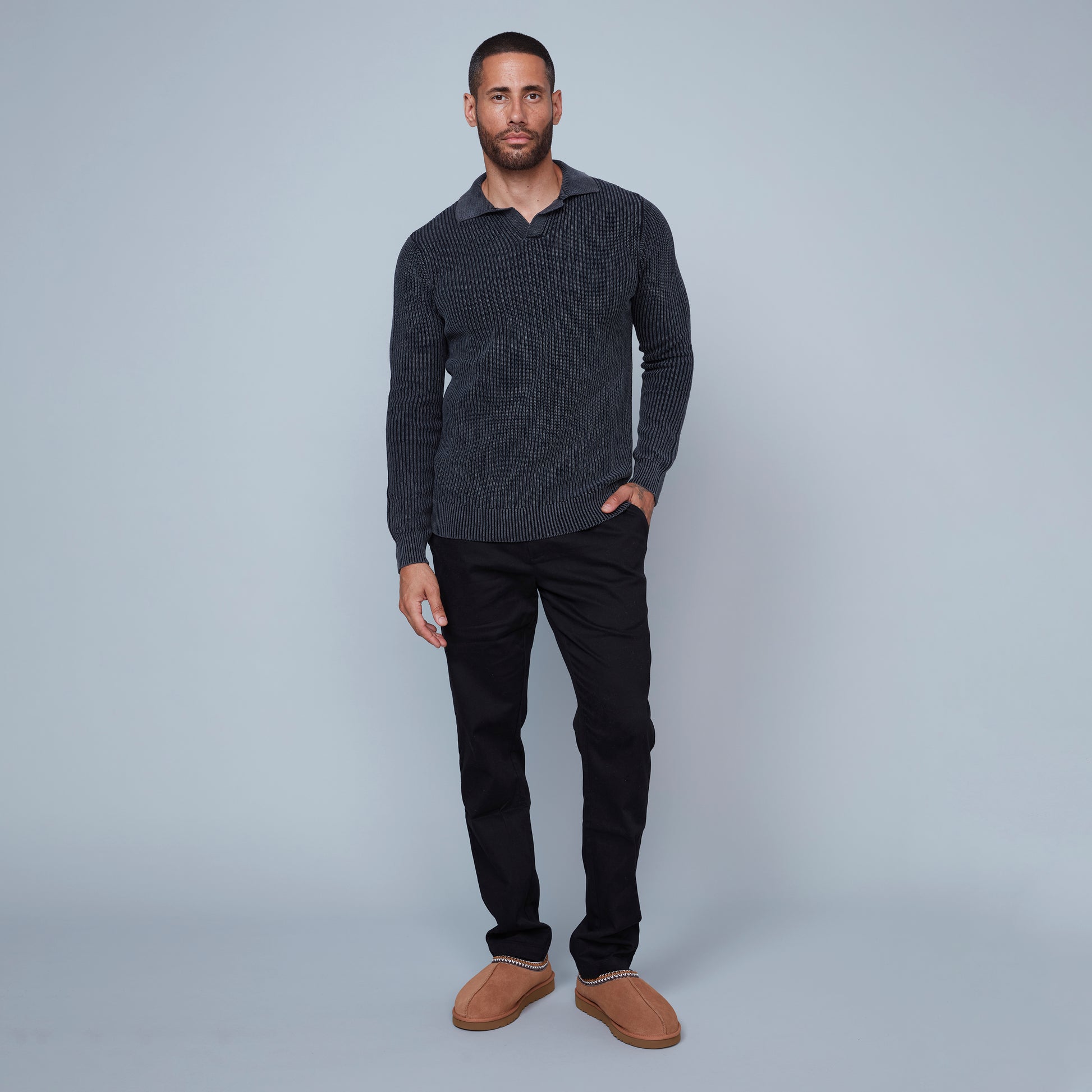 A man with short hair and a beard is standing against a plain light gray background. He is wearing a long-sleeve, textured, dark gray Johnny Collar Sweater from HEDGE, made from 100% cotton, paired with black pants. His left arm hangs by his side, revealing a tattoo on his hand and wrist, showcasing subtle fashion expertise.