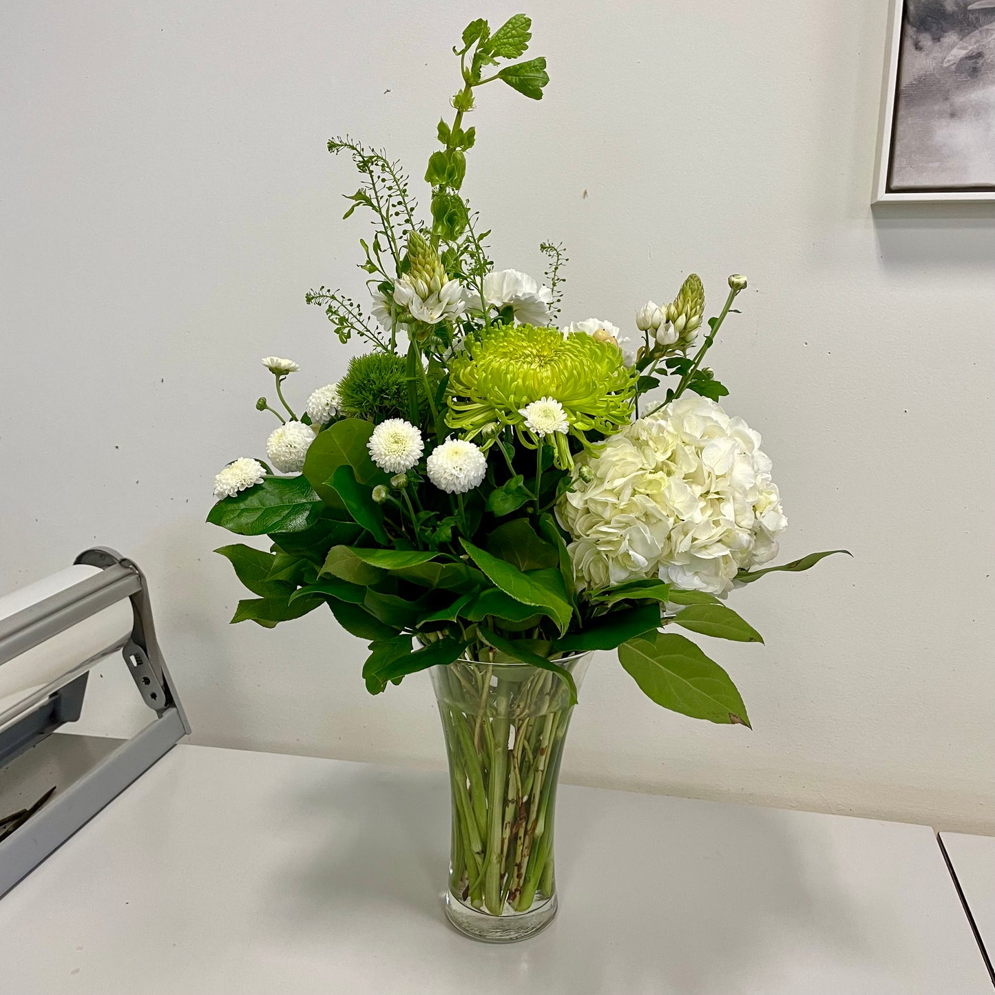 A Designer’s Choice Vase from Posies Flowers & Fashion, filled with white lilies and green foliage, graces a marble table in an elegantly modern room. Soft lighting and contemporary furniture in the background create a warm and inviting atmosphere, showcasing the artistry of floral designers, subject to availability.