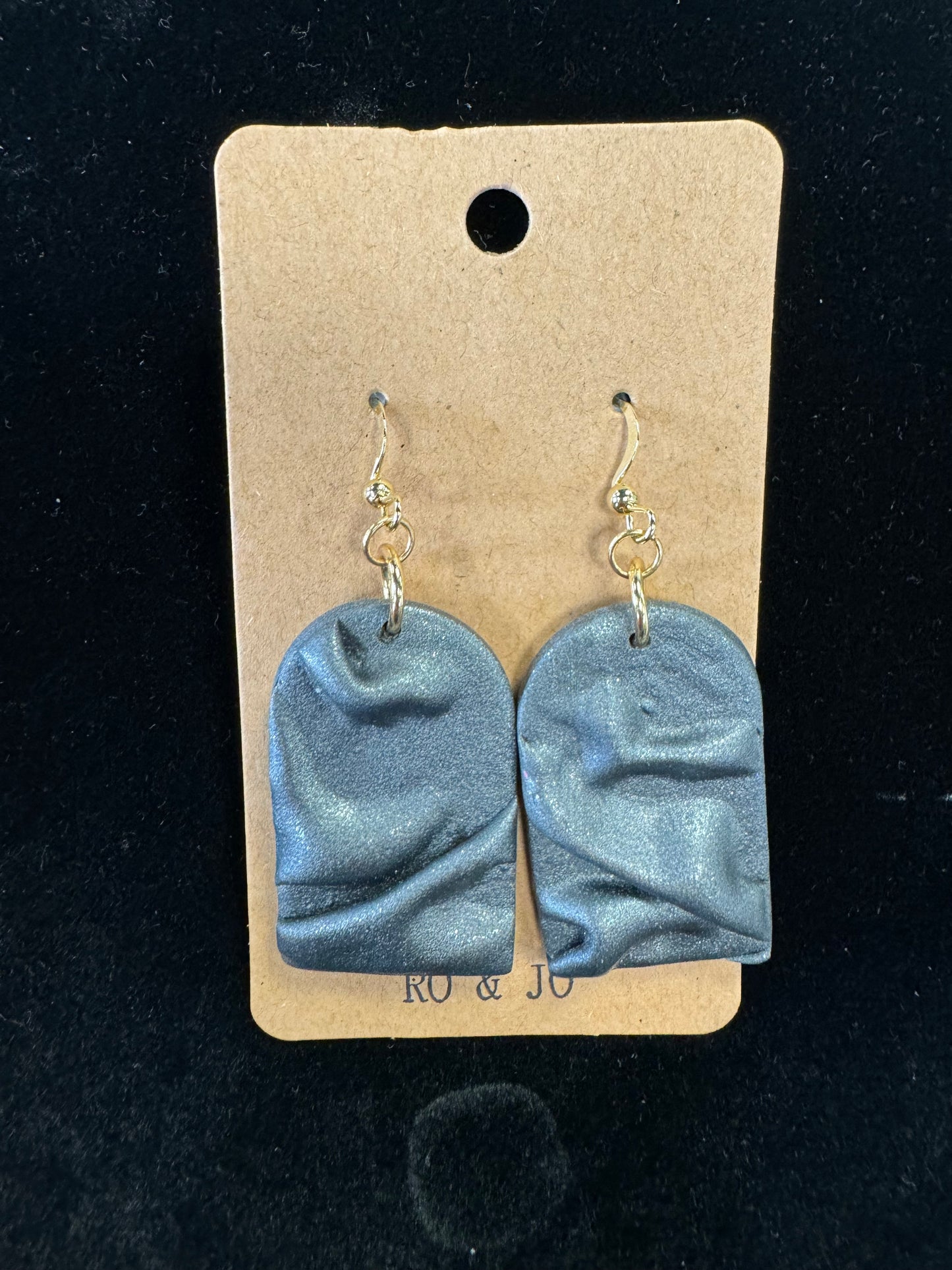 A pair of Ro&Jo Earrings 440, handmade from polymer clay with a textured, shiny gray finish, is displayed on a brown rectangular card with a small hole at the top. The RO&JO earrings feature uneven surfaces that create a wavy appearance and hang from gold hooks. The background is black.