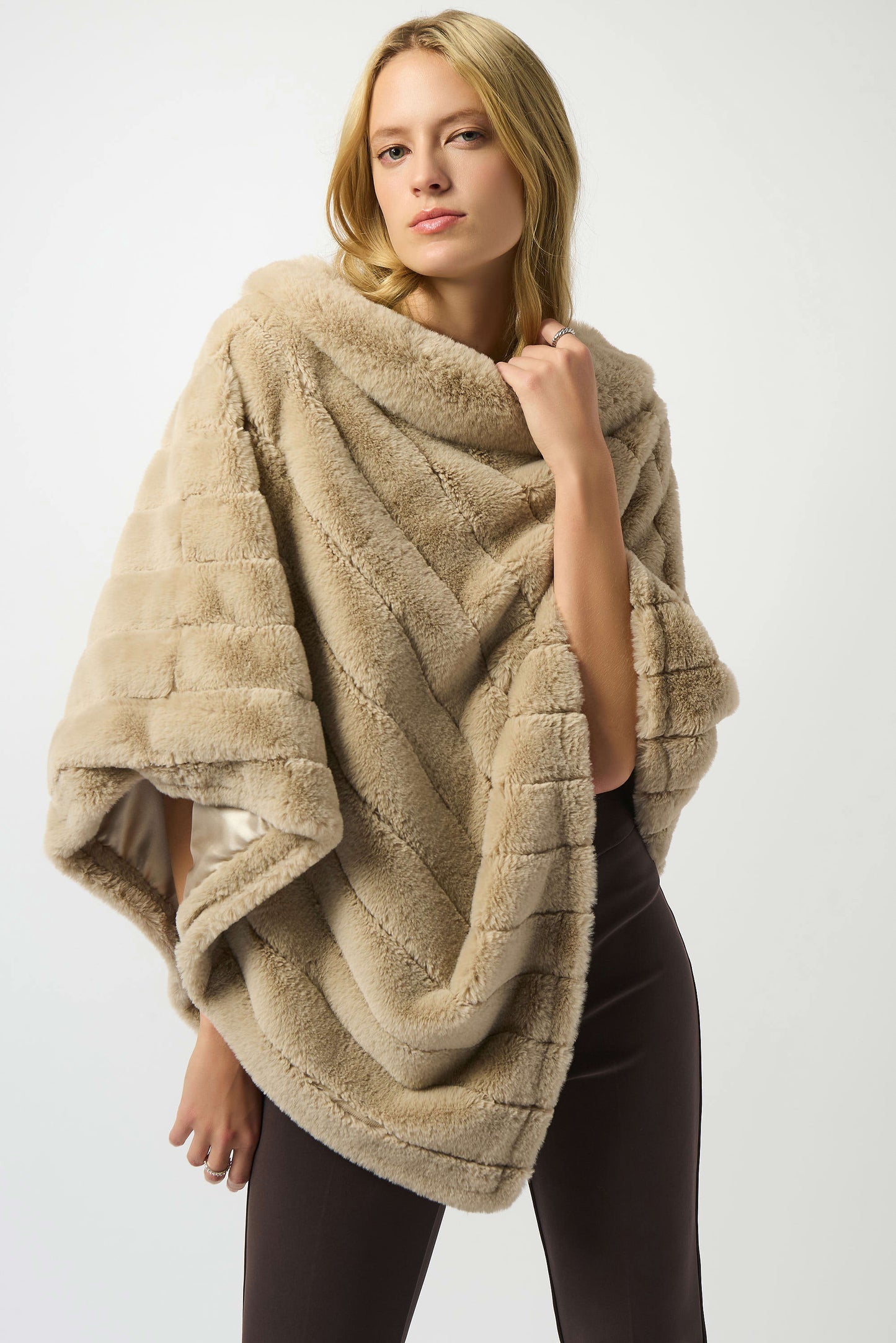 A woman with long blonde hair models the Joseph Ribkoff Faux Fur Textured Stripe Poncho 253932 over dark pants, standing confidently against a white background and holding the poncho closed.