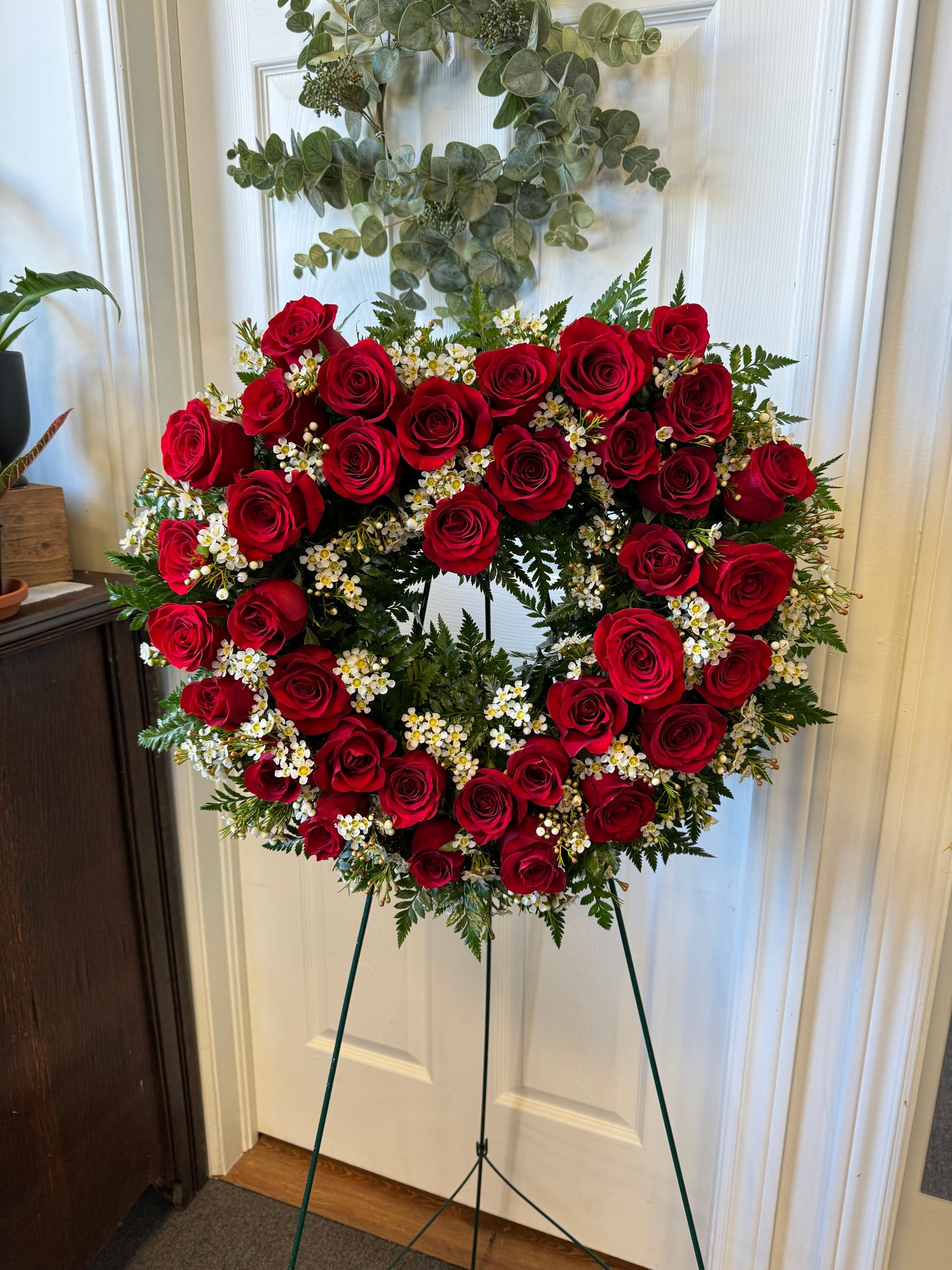 The Expression of Love wreath by Posies Flowers & Fashion, a heart-shaped arrangement featuring red roses and small white flowers accented with green foliage, is displayed on a green stand in front of a white door.