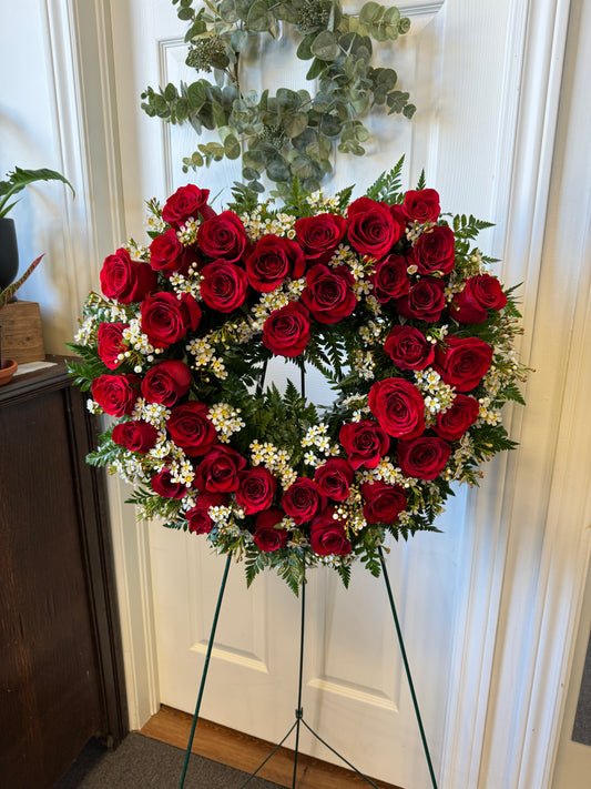 The Expression of Love wreath by Posies Flowers & Fashion, a heart-shaped arrangement featuring red roses and small white flowers accented with green foliage, is displayed on a green stand in front of a white door.