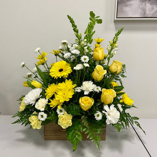 The "Sunshine Tribute" from Posies Flowers & Fashion is a wooden box brimming with a lively floral arrangement that includes yellow roses, yellow gerbera daisies, white carnations, white chrysanthemums, and green foliage. The flowers are beautifully arranged to offer a fresh and cheerful display against a neutral background.