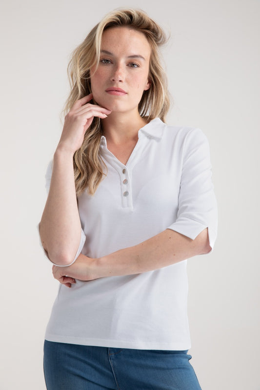 A person with long hair in a 100% cotton white collared shirt, Top Polo 7838 - MARBLE by MARBLE, poses thoughtfully, hand on chin. They're against a plain marble-like backdrop, wearing blue pants that reflect the classic style of the brand.