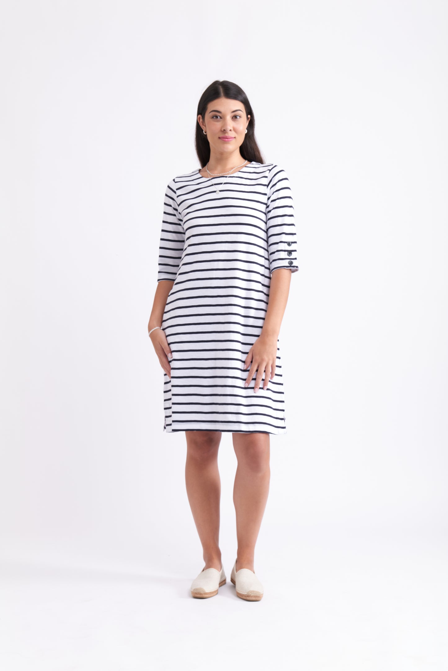 A woman stands against a white background wearing the FOIL Oh Buoy Dress FO7952, a 100% cotton, knee-length summer dress with navy blue horizontal stripes and elbow-length sleeves. She pairs it with beige slip-on shoes and has straight, dark hair.