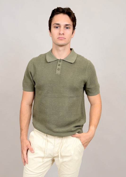 Dressed in a Knit Polo Light Olive 56MS372S by HEDGE, the individual stands against a beige background. With their right hand in their pocket and a neutral expression, the 100% cotton knit of the short-sleeved top enhances their casual elegance alongside light-colored pants.