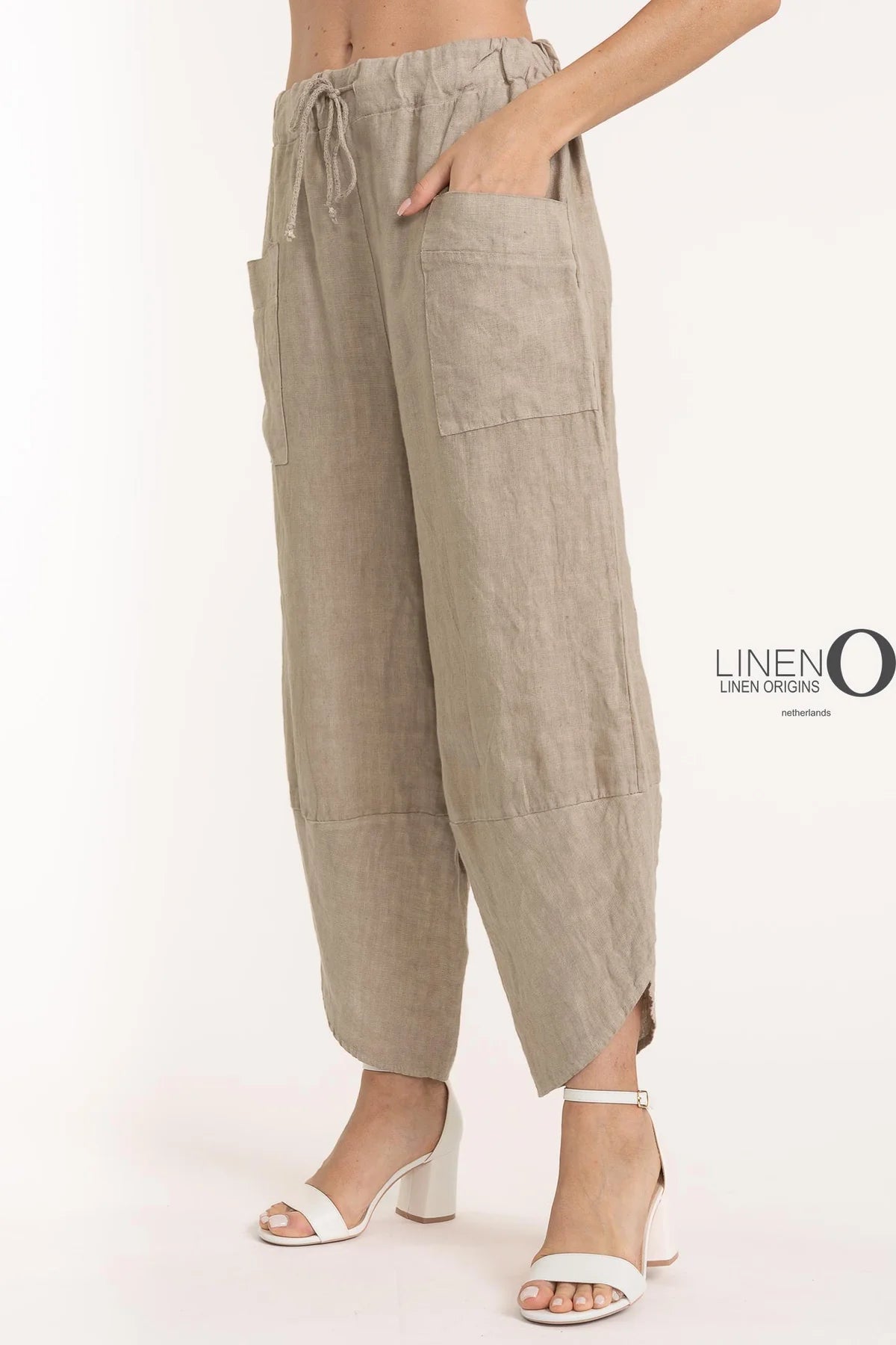Someone wears LinenO's French Linen Pant PT841-148 Natural, featuring wide legs, large front pockets, and a drawstring waist. They pair it with an eco-friendly French linen blouse and white strappy high-heeled sandals against a plain off-white background.