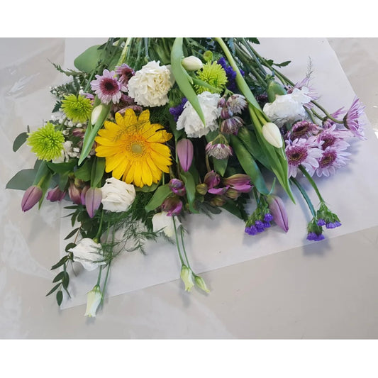 A vibrant bouquet labeled "Spring Bound" from Posies Flowers & Fashion adorns a vase on a flat surface. The arrangement features yellow gerbera daisies, white roses, purple tulips, green chrysanthemums, and an assortment of other flowers mixed with diverse greenery. This fresh and colorful bouquet showcases natural beauty impeccably.