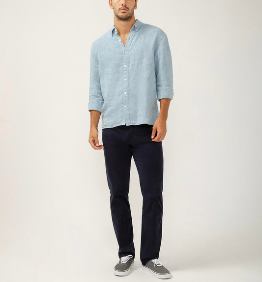 A man poses against a plain backdrop, wearing a light blue button-up and Machray Athletic Fit Straight Leg Pants Navy by Silver Jeans Co. His slightly wrinkled shirt enhances the relaxed stance, while gray sneakers finish his effortlessly stylish outfit.