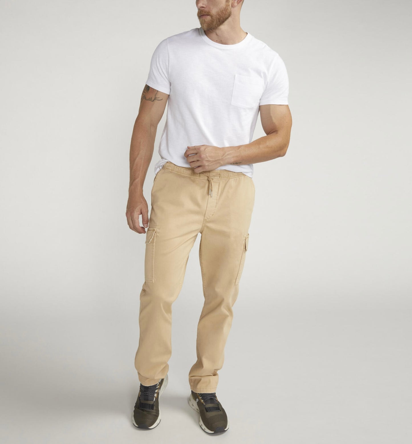 A man in a white t-shirt and Silver Jeans Co.'s Pull-On Cargo Essential Twill Pant with an adjustable waistband stands against a plain background. He's sporting black sneakers, showcases a tattoo on his left arm, and casually tucks his right hand into his pocket.