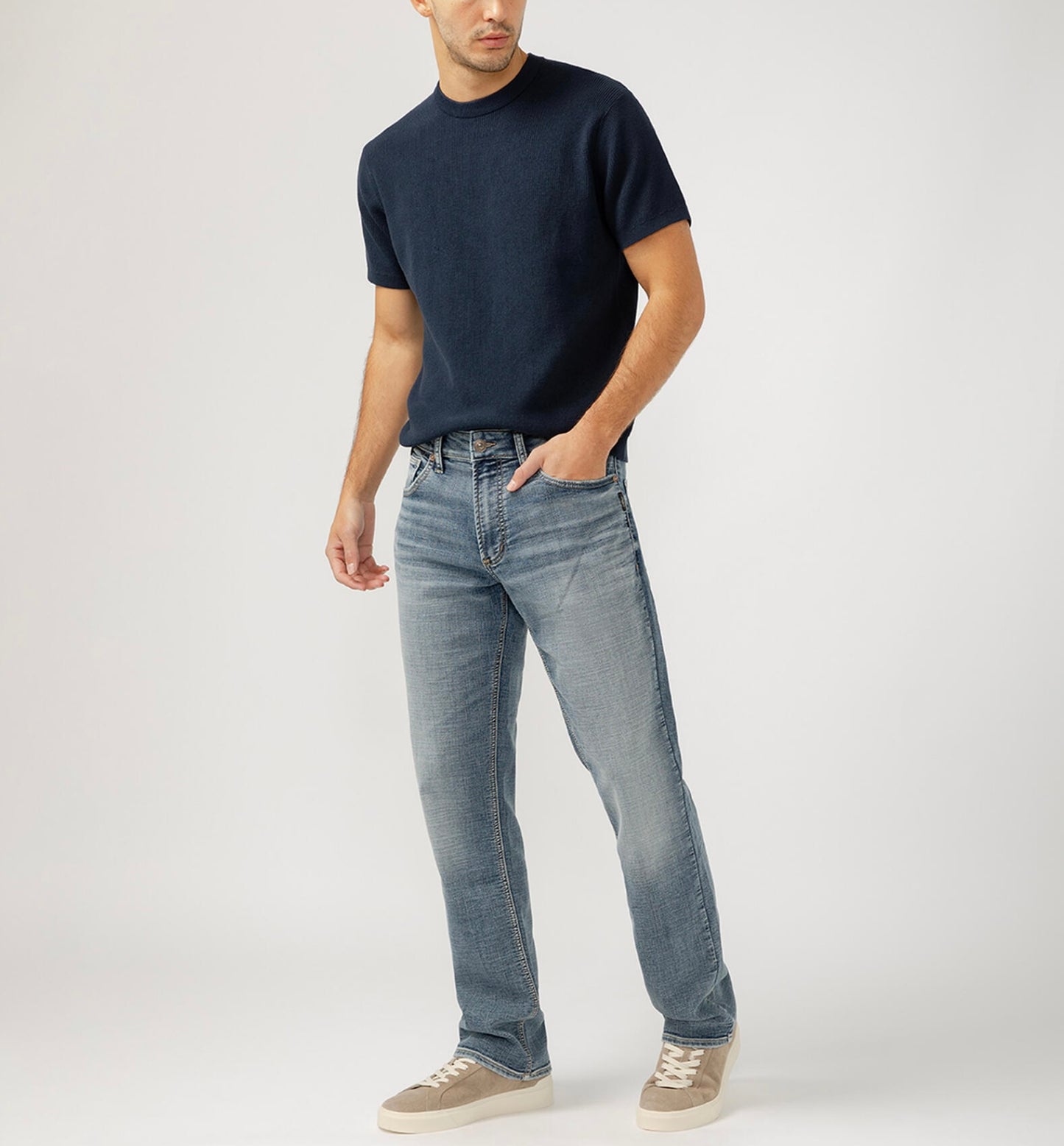 Dressed in a black t-shirt and Silver Jeans Co.'s Grayson Classic Fit Straight Leg Jeans, a person stands casually with a hand in their pocket. They sport gray sneakers against a plain light gray backdrop.