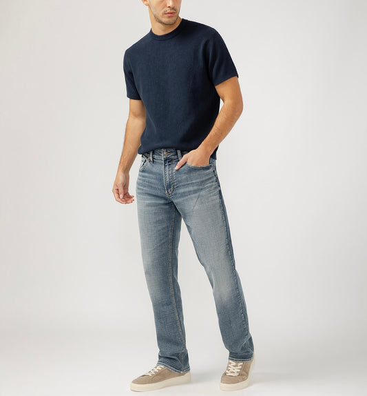 Dressed in a black t-shirt and Silver Jeans Co.'s Grayson Classic Fit Straight Leg Jeans, a person stands casually with a hand in their pocket. They sport gray sneakers against a plain light gray backdrop.