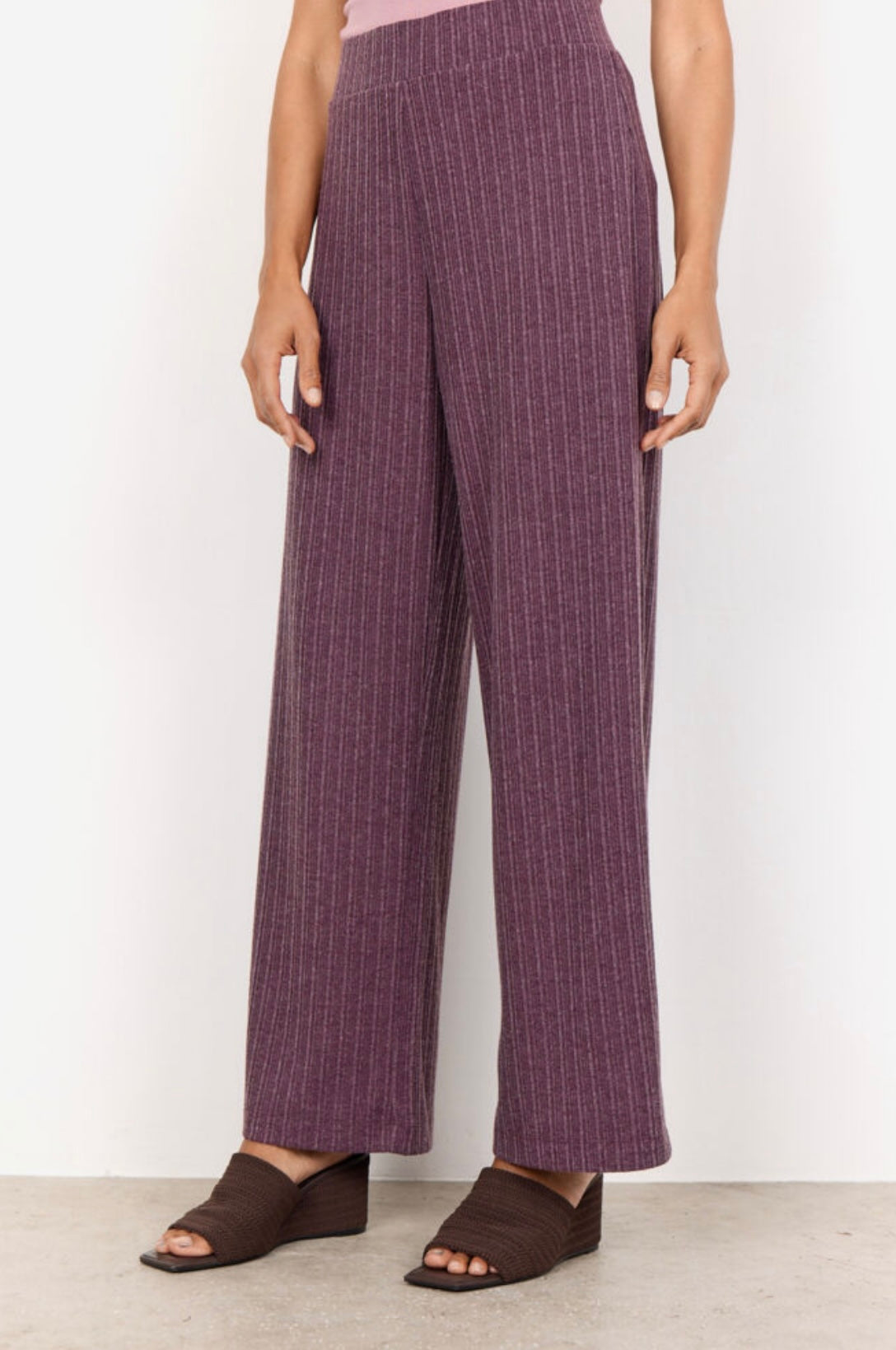 A person with short curly hair is standing against a plain white background. They are wearing a sleeveless pink top and the SC-ISEL 1 Pants by SoyaConcept, which are high-waisted, wide-leg purple pants featuring a subtle pinstripe design. Made from recycled polyester, the pants complement their arms slightly crossed as they look at the camera.