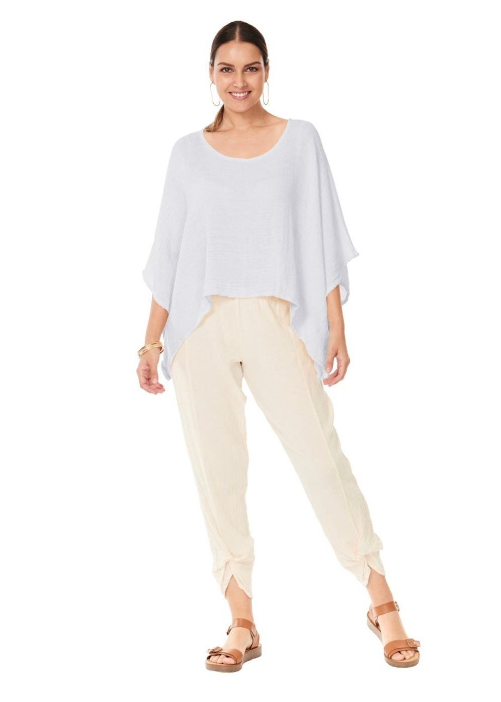A woman stands smiling against a plain white background, wearing the HAVANA SNOW TOP by Oh My Gauze, which is an asymmetrical sleeveless gauze top. She pairs it with beige tied-ankle pants and accessorizes with large hoop earrings, bangles, and tan sandals with thick straps. Her dark hair is pulled back, completing this flattering design.