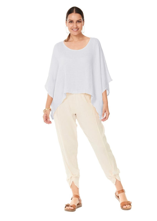 A woman stands smiling against a plain white background, wearing the HAVANA SNOW TOP by Oh My Gauze, which is an asymmetrical sleeveless gauze top. She pairs it with beige tied-ankle pants and accessorizes with large hoop earrings, bangles, and tan sandals with thick straps. Her dark hair is pulled back, completing this flattering design.