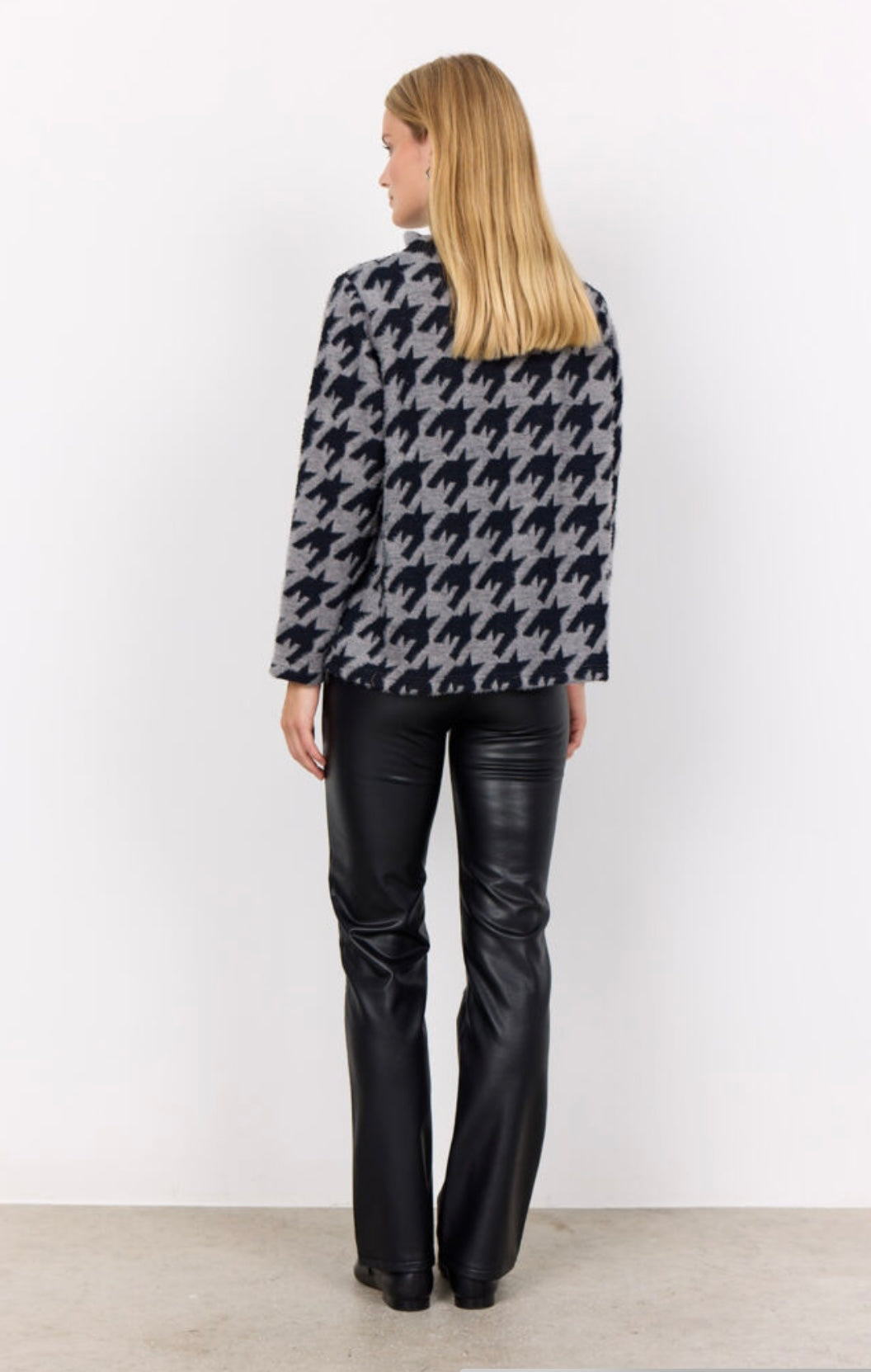 A person with long hair is wearing the SC-IRIT 1 Top from SoyaConcept, a high-neck, long-sleeve sweater adorned with a large black and grey houndstooth pattern. Over it, they are clad in a loose fit feminine waistcoat made of cotton blend. They also have on fitted black pants and are standing against a plain white background, looking straight at the camera.