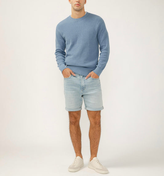 A person is in a minimal and bright setting, wearing a light blue sweater, Silver Jeans Co.'s Classic Fit Luxe Heritage Everyday Short M53150EFS184. Their hands are in their pockets, and they're sporting white sneakers against a plain background.