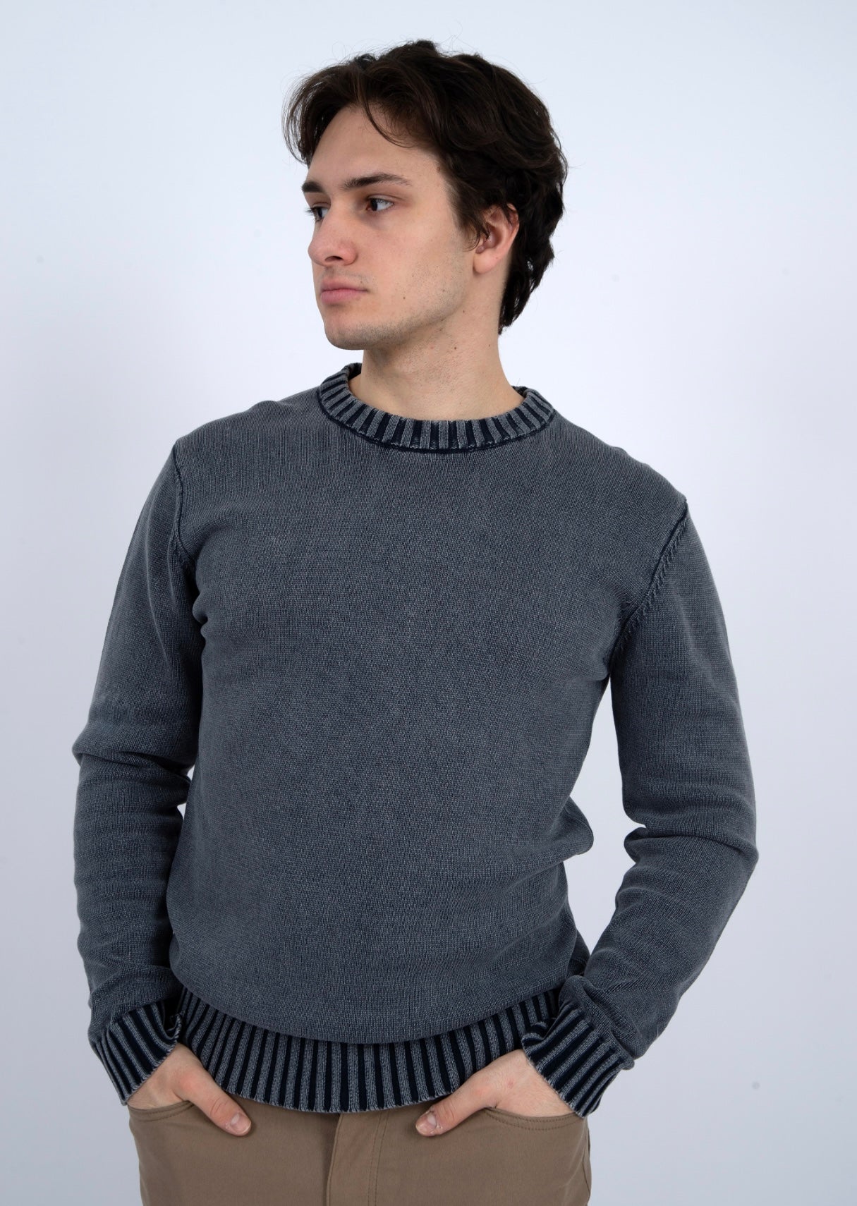 A young man with medium-length brown hair wears the HEDGE CREW SWEATER-NAVY ACID WASH, featuring striped cuffs and collar, standing with hands in pockets and looking to the side against a plain light background.