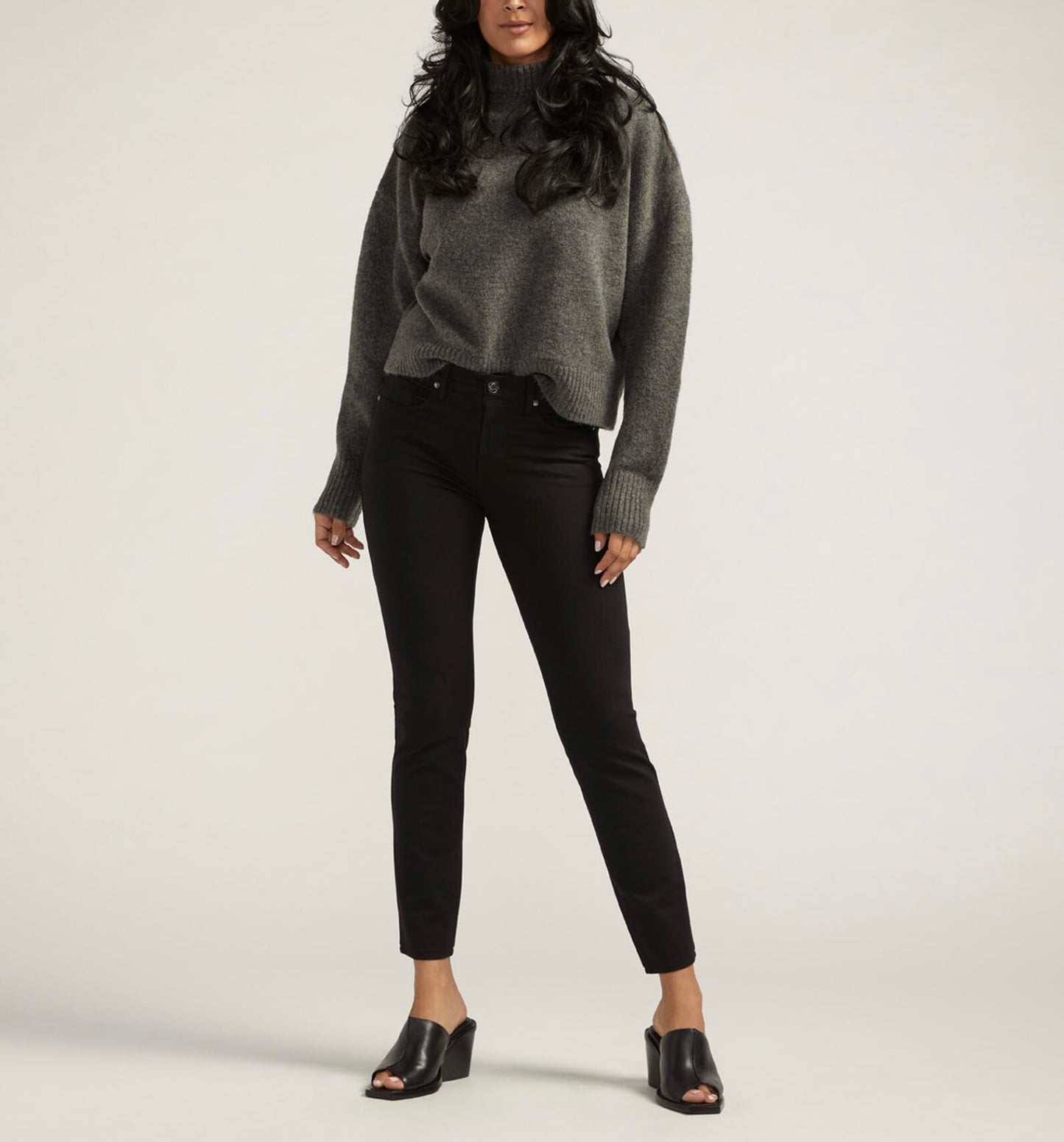 A person stands against a neutral background wearing an oversized gray sweater, JAG's Cassie Mid Rise Slim Straight Ponte Pant in black, and black open-toe heeled mules. Their hands hang relaxed at their sides, and their long hair cascades in waves.