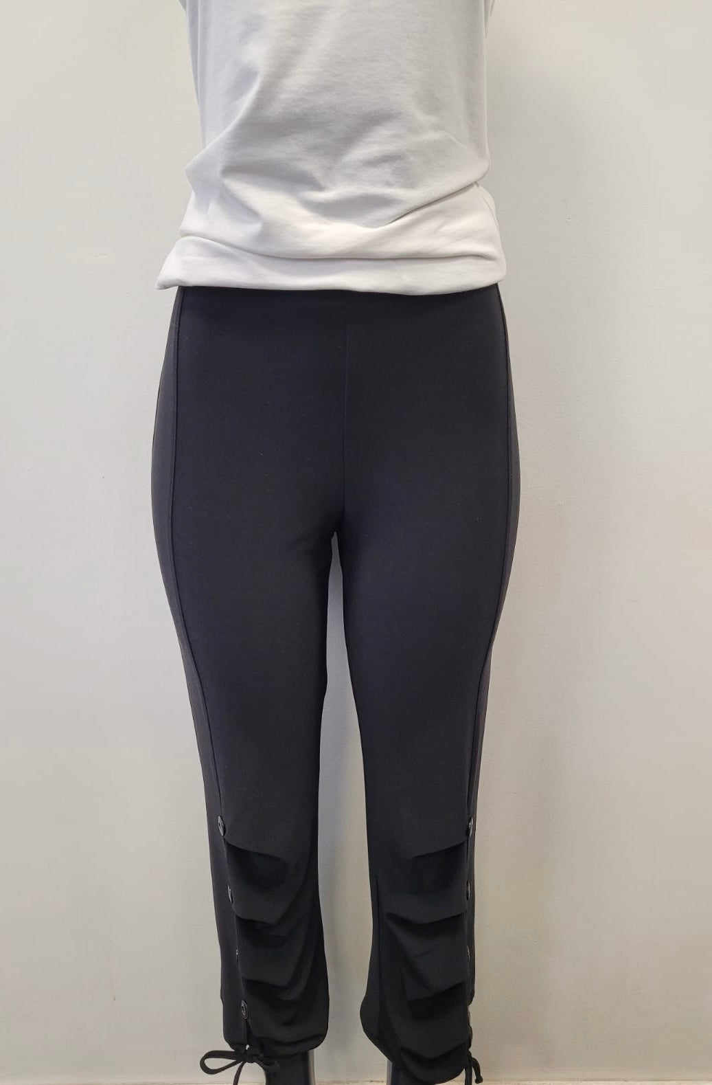 A person models Bali Corp’s Black Leggings 6279-Bali Corp, featuring decorative button and tie details near the ankles, paired with a white t-shirt, against a plain light-colored wall. Made in Canada.