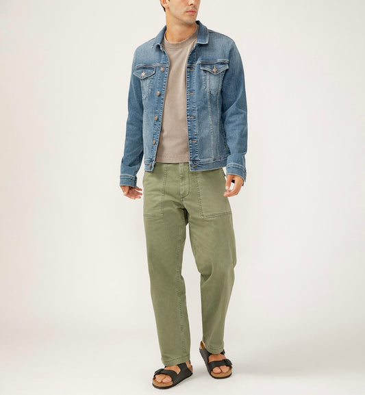 A person in a Silver Jeans Co. Denim Jacket MJ0014EAD309 - medium indigo wash over a beige shirt, green pants, and black sandals stands against a plain background, looking to the side with relaxed hands.