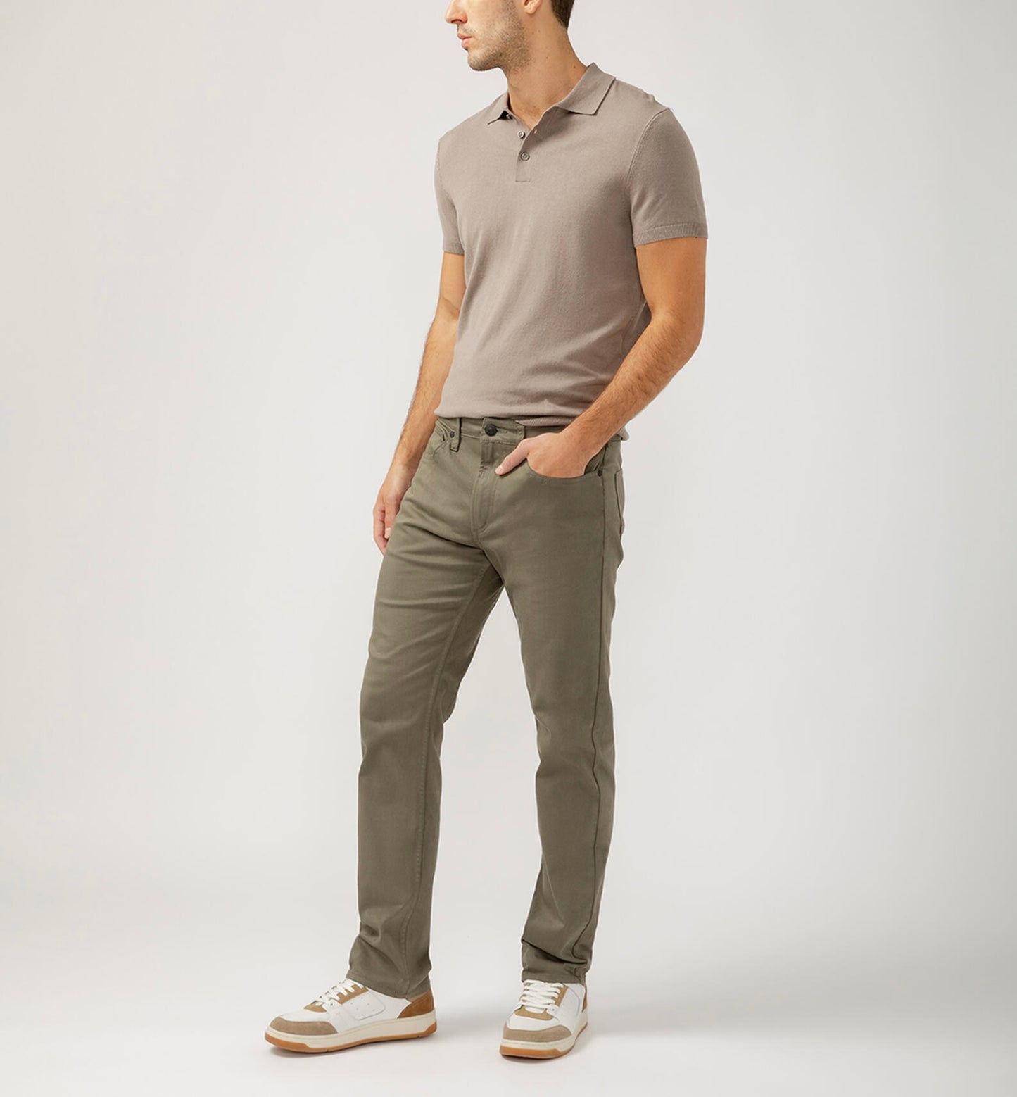 A person wearing a beige polo, Machray Athletic Fit Straight Leg Pants M63410LBS659 by Silver Jeans Co. in olive green, and white sneakers with brown accents stands against a light gray background, posing with one hand in their pocket and looking left.