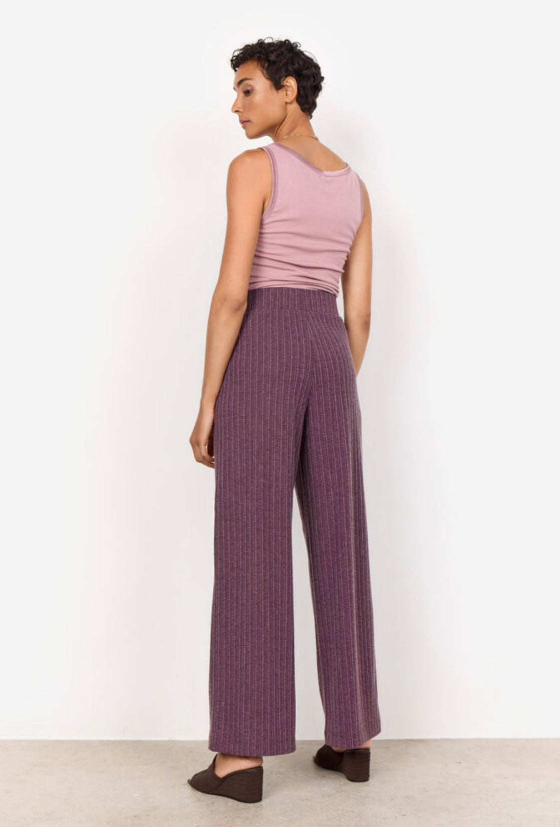 A person with short curly hair is standing against a plain white background. They are wearing a sleeveless pink top and the SC-ISEL 1 Pants by SoyaConcept, which are high-waisted, wide-leg purple pants featuring a subtle pinstripe design. Made from recycled polyester, the pants complement their arms slightly crossed as they look at the camera.