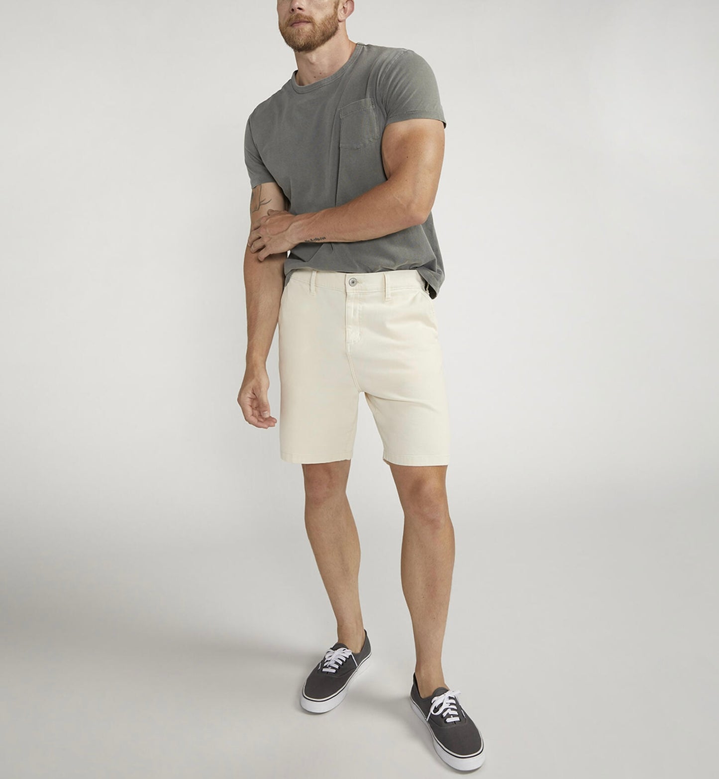 A person wearing a gray T-shirt, Silver Jeans Co.'s Painter Short M53152CTL640 in classic fit, and black sneakers with white soles stands against a plain gray background with their right hand on their left arm.