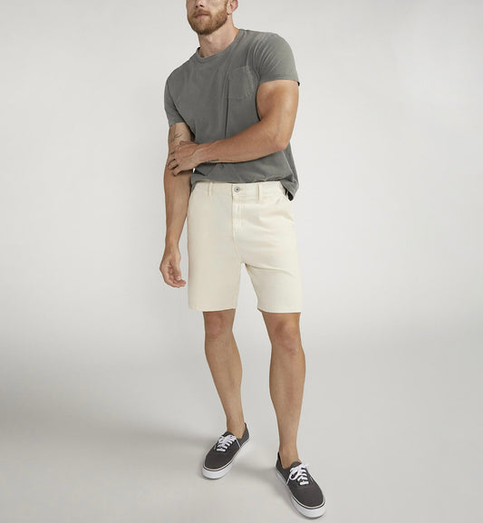 A person wearing a gray T-shirt, Silver Jeans Co.'s Painter Short M53152CTL640 in classic fit, and black sneakers with white soles stands against a plain gray background with their right hand on their left arm.