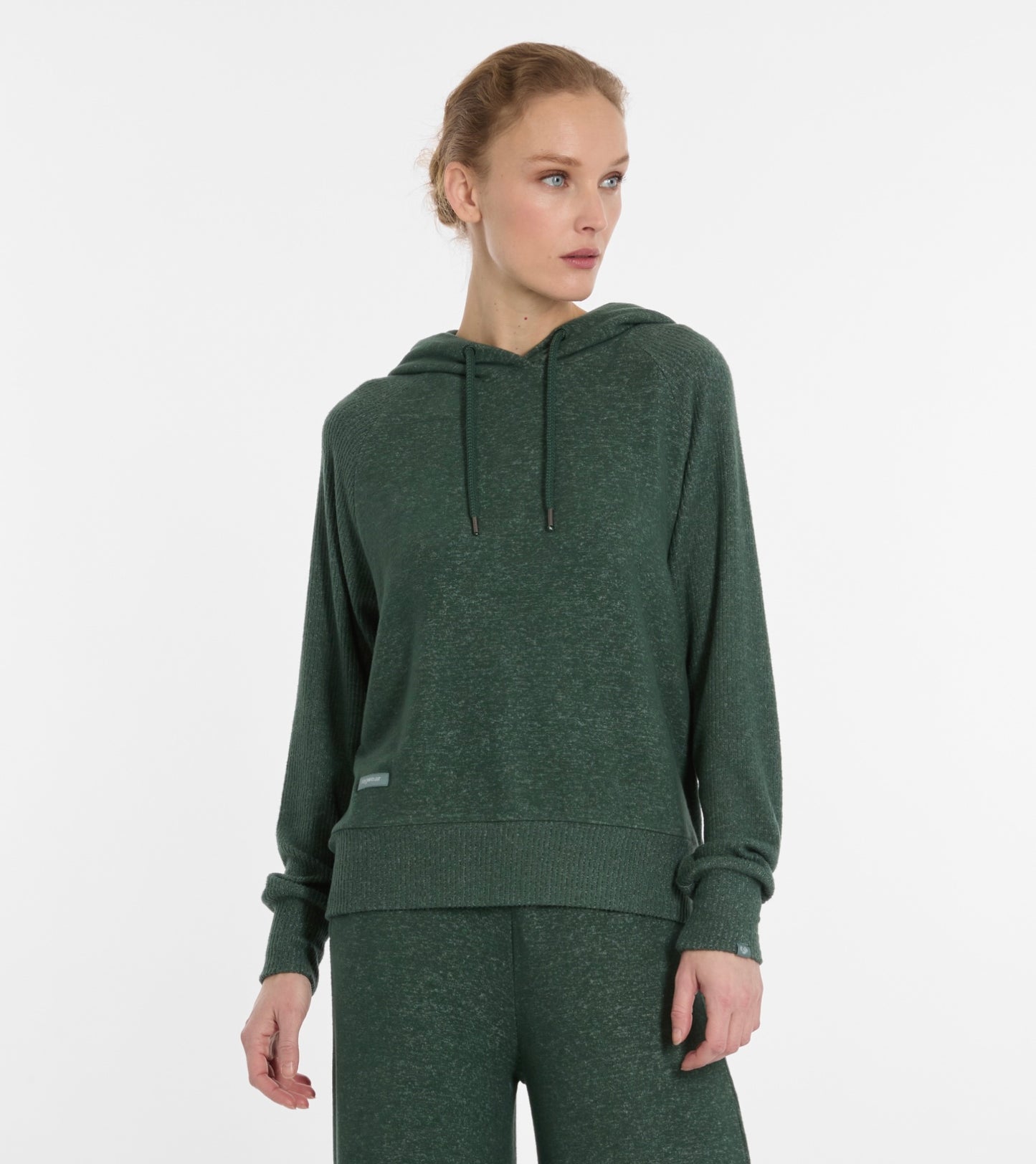 A woman in a pine green TONNARA Women’s Sweatshirt by RAGWEAR with matching pants stands against a plain white background, looking slightly to her left.