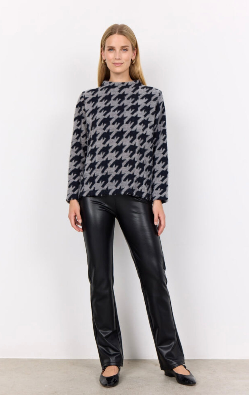A person with long hair is wearing the SC-IRIT 1 Top from SoyaConcept, a high-neck, long-sleeve sweater adorned with a large black and grey houndstooth pattern. Over it, they are clad in a loose fit feminine waistcoat made of cotton blend. They also have on fitted black pants and are standing against a plain white background, looking straight at the camera.