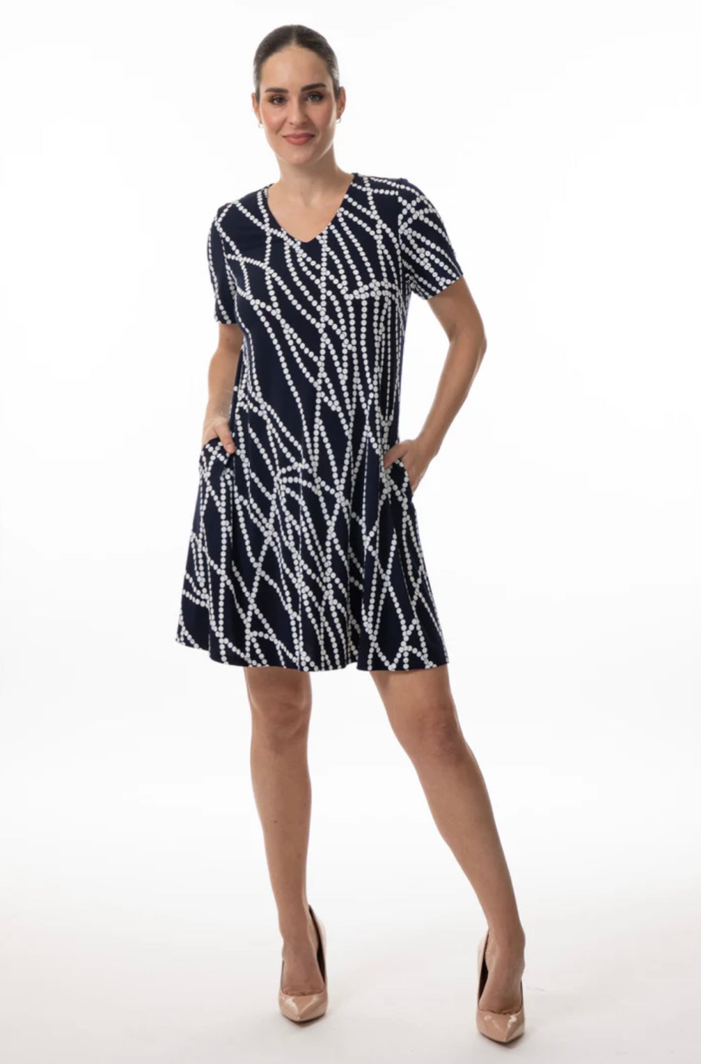 A person stands confidently in Bali Corp's Dress 8635, a navy polyester piece with a white abstract line pattern, featuring short sleeves and pockets. They smile in nude heels against a plain white backdrop, exuding style.