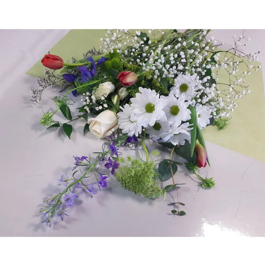 A vibrant floral arrangement known as Spring Dew from Posies Flowers & Fashion lays on a light-colored surface, displayed in a delicate vase. The bouquet includes white daisies, baby's breath, lavender and purple flowers, and a mix of white, red, and peach roses, all accented with green foliage. Availability may vary due to seasonal flowers. The background features a pale green paper.