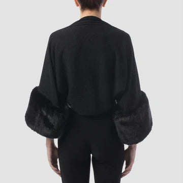 A person is modeling the Cover Up Style 153483X - BLACK by Joseph Ribkoff, featuring an open front that reveals a fitted white top underneath and black pants. The black wrap's faux-fur cuffs stand out, adding a luxurious touch against a plain, light gray background.