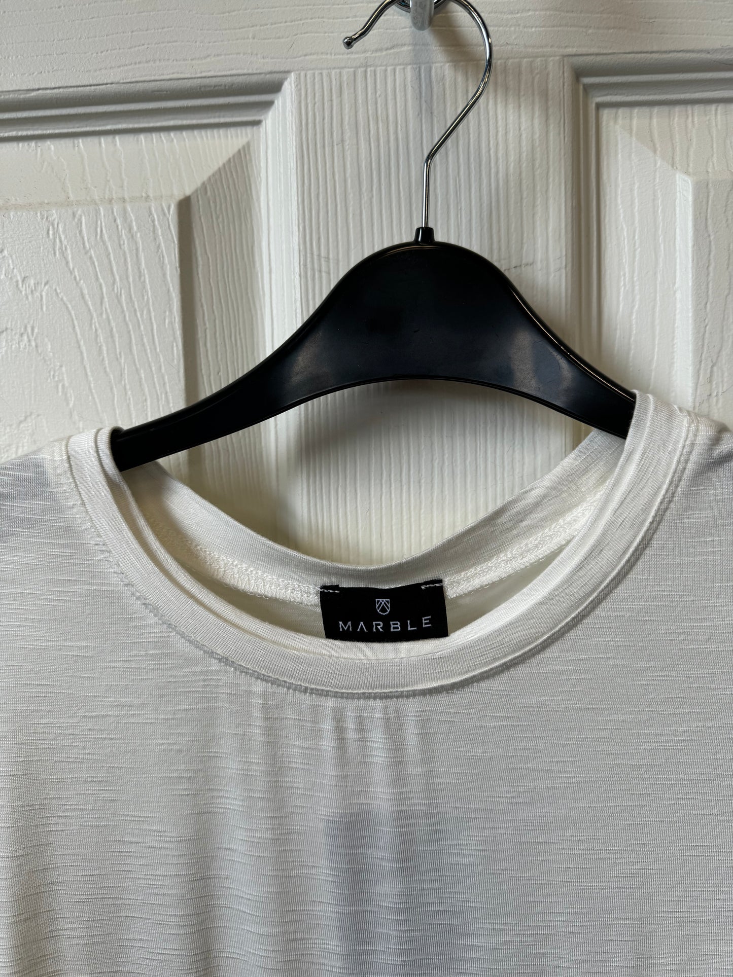 The Marble L/S Crewneck Top w/Ruche Cuff & Button Detail from MARBLE, featuring a round neckline and a long-sleeve design, is displayed on a black hanger against a white door. Made from a lightweight viscose blend, this white top showcases slightly sheer, textured fabric and ruched cuffs adorned with four small buttons. A black tag with white text is visible at the back of the neck.