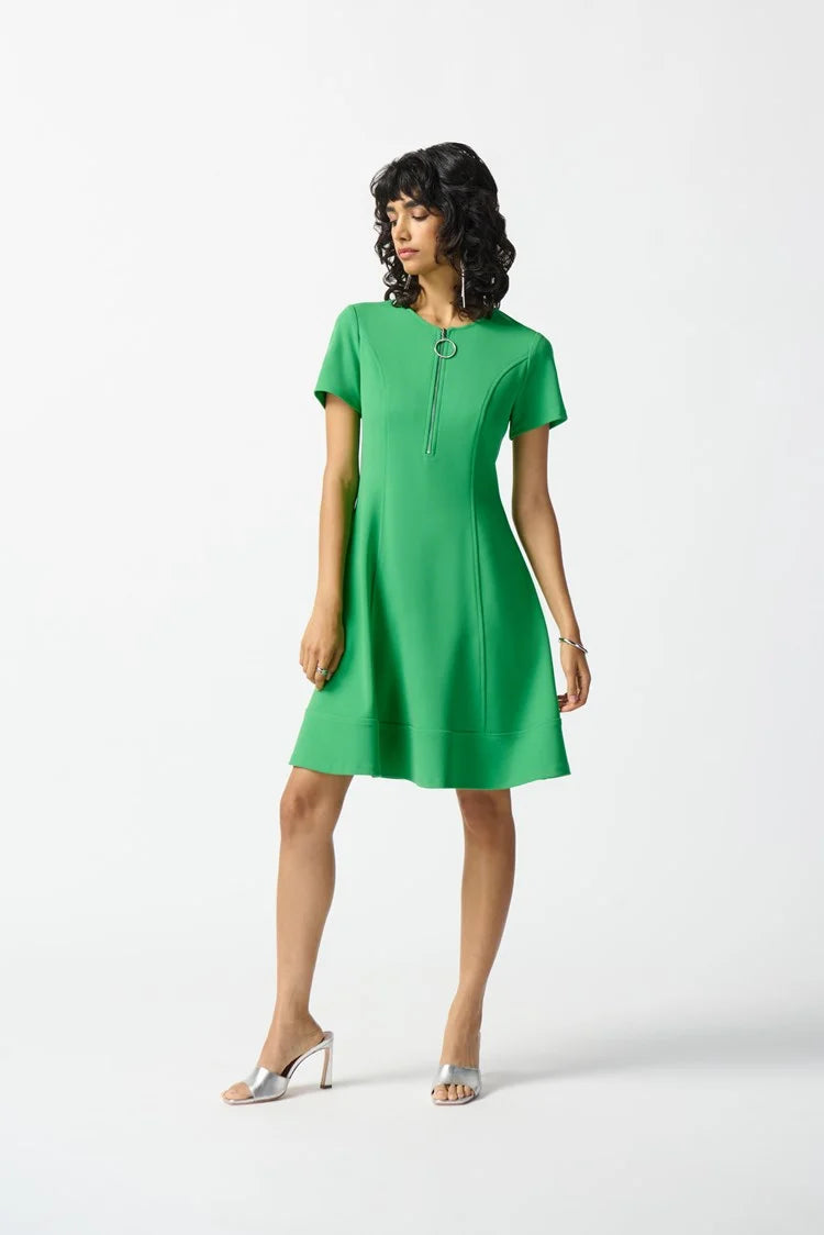 A person with shoulder-length dark wavy hair stands against a plain white background, wearing the Joseph Ribkoff - 242031 Island Green Scuba Crepe Fit and Flare Dress. The short-sleeved attire, featuring a zipped neckline and showcasing a fit and flare silhouette, is accessorized with a silver bracelet and white heeled sandals.