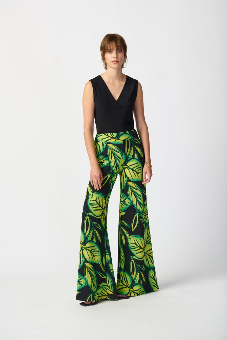A person with short brown hair is wearing a Silky Knit Sleeveless Top 241239 by Joseph Ribkoff in black, along with large hoop earrings. They are also sporting a bracelet and a skirt featuring a green leaf pattern. The background is plain white.