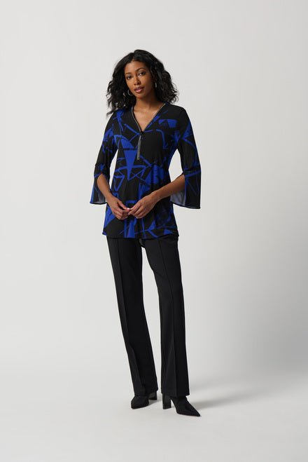 A woman with shoulder-length wavy black hair is wearing a Joseph Ribkoff Three-quarter Sleeve Tunic Style 234279, which features an abstract blue pattern. She is gazing slightly off to the side with a neutral expression. The tunic, crafted from colorful fabric, showcases a prominent zipped V-neckline. The background is plain and light-colored.