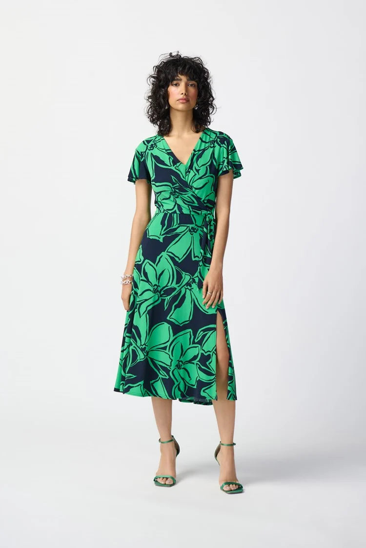 A woman with curly blonde hair, wearing a green Joseph Ribkoff Floral Print Silky Knit Flowy Wrap Dress with ruffled sleeves, sits barefoot on the edge of a stone fountain in a sunlit courtyard. She gazes thoughtfully at the water beside her. Lush greenery and architectural details fill the background.