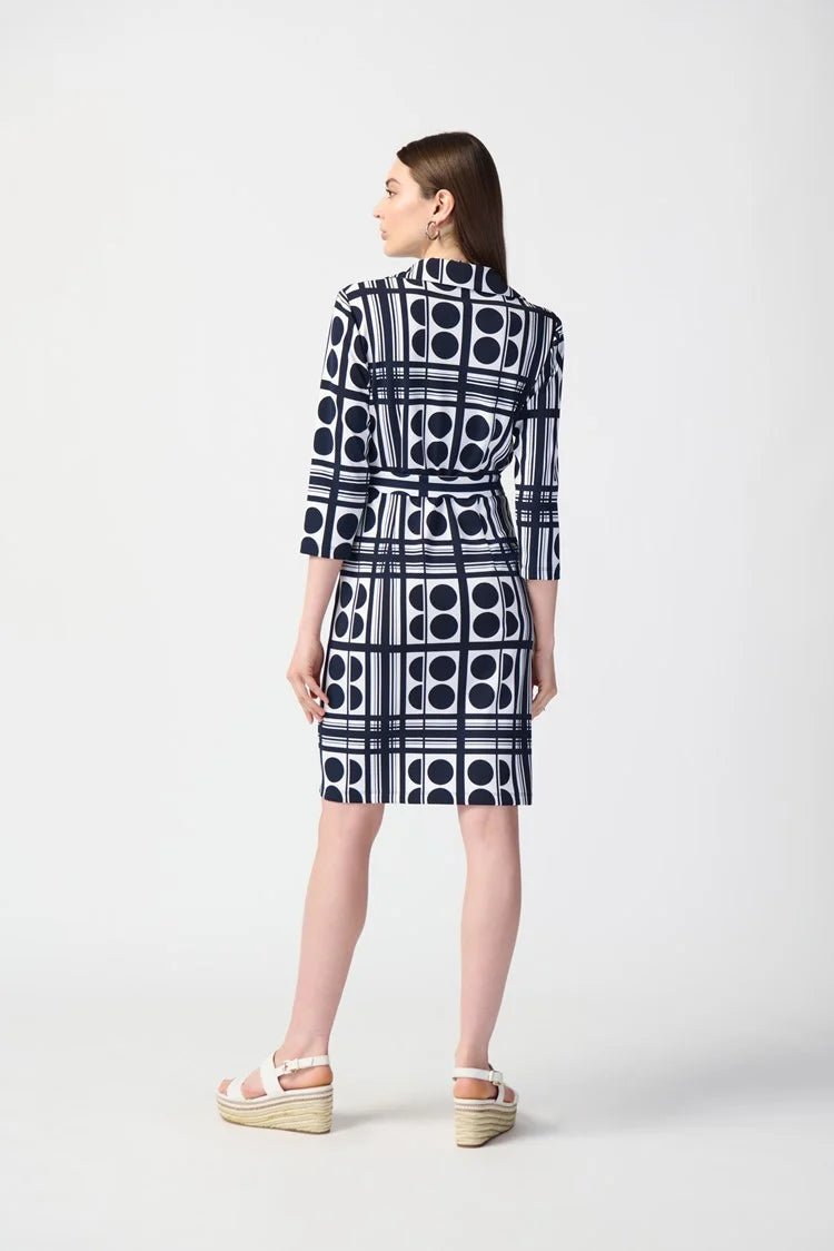 A woman stands wearing the Joseph Ribkoff Dot Print Silky Knit Belted Shirt Dress 241001, which is knee-length with a navy and white geometric pattern. The dress features three-quarter sleeves and includes a matching waist tie that creates a flattering silhouette. She has long, straight brown hair and is paired with white platform sandals. The background is plain white.