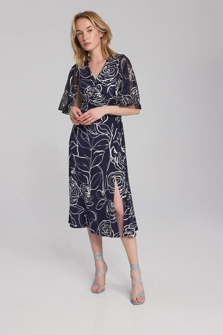A blonde woman is posing in a Floral Print Silky Knit and Chiffon Dress 241764 from Joseph Ribkoff. The dress, part of the Signature Collection, features short sheer cape sleeves and a knee-length hem. She is seated with one hand on her knee and the other on her hip, wearing light blue strappy high-heeled sandals.