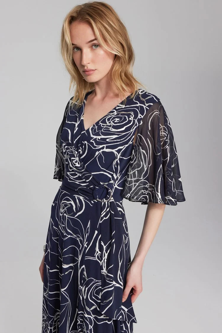 A blonde woman is posing in a Floral Print Silky Knit and Chiffon Dress 241764 from Joseph Ribkoff. The dress, part of the Signature Collection, features short sheer cape sleeves and a knee-length hem. She is seated with one hand on her knee and the other on her hip, wearing light blue strappy high-heeled sandals.