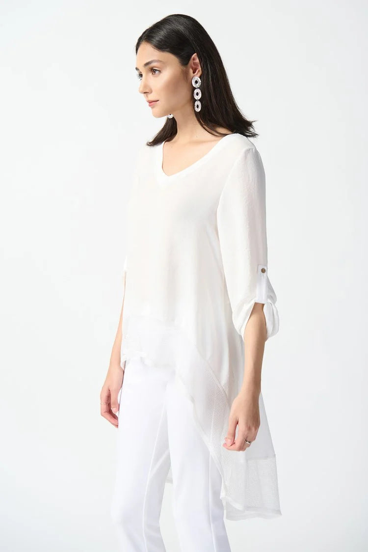A woman stands against a plain white background wearing the Gauze and Mesh High Low Flare Top 242066 Joseph Ribkoff, a flowy, semi-sheer tunic by Joseph Ribkoff in gauze fabric with three-quarter sleeves that button at the cuffs. She pairs the top with fitted white pants and white open-toe heels, accessorizing with long, dangling earrings.