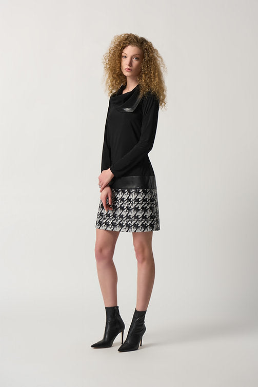 A woman with curly hair stands in a light-filled room wearing a Joseph Ribkoff Houndstooth Cowl Neck Dress Style 233130 and black ankle boots. Behind her, there is a mustard-yellow couch against a wall adorned with white decorative molding.