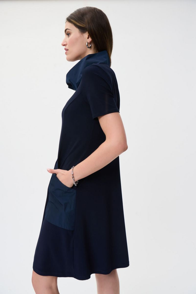 A woman stands against a plain white background, wearing the Drawstring Shirt Dress Style 231141 by Joseph Ribkoff. This navy blue shirt dress is crafted from lightweight fabric and features side pockets and a drawstring collar. She has long, dark hair and is accessorized with subtle jewelry, including a bracelet and earrings. One of her hands rests near her neck.