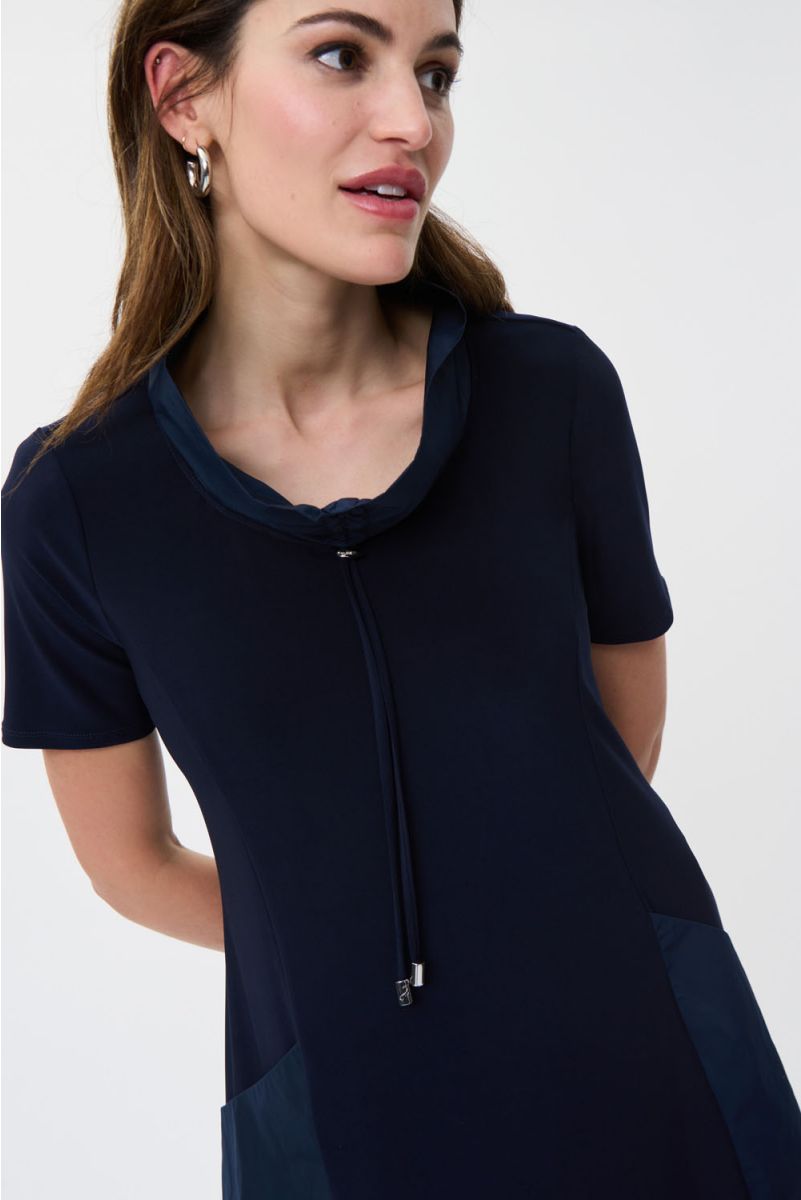A woman stands against a plain white background, wearing the Drawstring Shirt Dress Style 231141 by Joseph Ribkoff. This navy blue shirt dress is crafted from lightweight fabric and features side pockets and a drawstring collar. She has long, dark hair and is accessorized with subtle jewelry, including a bracelet and earrings. One of her hands rests near her neck.
