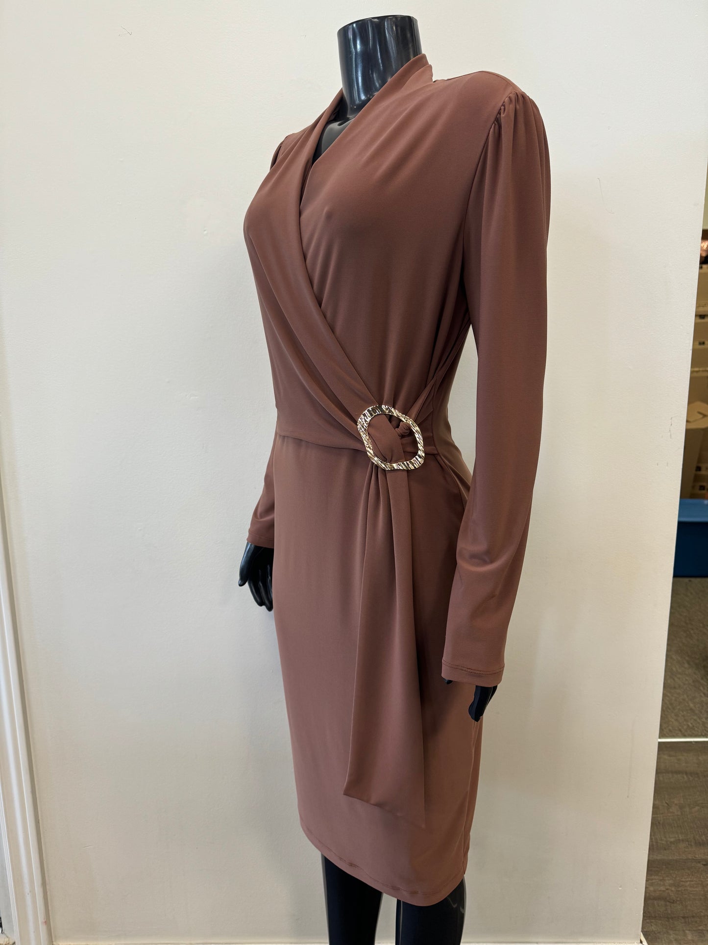 A mannequin displays the Wrap Front Belted Dress Style 233119 by Joseph Ribkoff, an elegant long-sleeved, knee-length brown fall dress featuring a deep V-neckline. The dress has a side buckle detail on the waist that creates a gathered effect. It is set against a plain, off-white background.