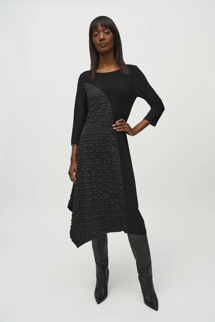 A person with long, straight hair is confidently posing with arms crossed against a plain background. They are wearing the Silky Knit And Jacquard Handkerchief Dress by Joseph Ribkoff, which features an asymmetrical cut and textured pattern on one side. They accessorize with silver bracelets on one wrist.