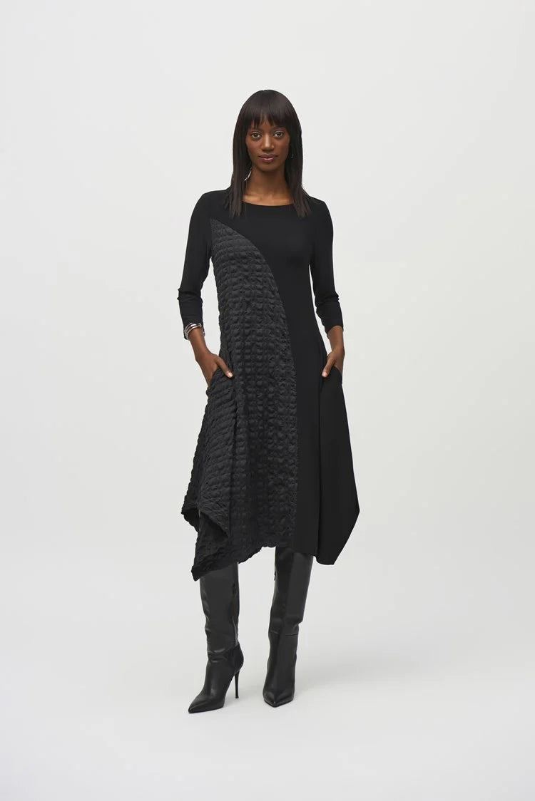 A person with long, straight hair is confidently posing with arms crossed against a plain background. They are wearing the Silky Knit And Jacquard Handkerchief Dress by Joseph Ribkoff, which features an asymmetrical cut and textured pattern on one side. They accessorize with silver bracelets on one wrist.