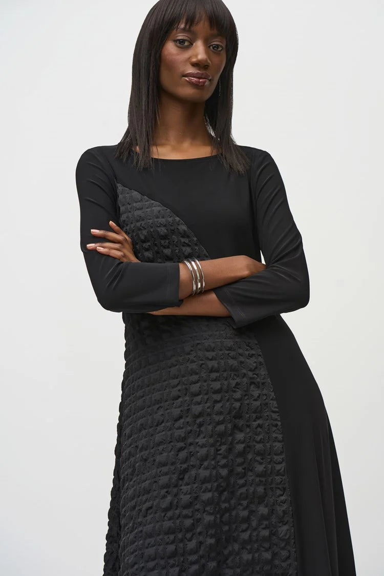 A person with long, straight hair is confidently posing with arms crossed against a plain background. They are wearing the Silky Knit And Jacquard Handkerchief Dress by Joseph Ribkoff, which features an asymmetrical cut and textured pattern on one side. They accessorize with silver bracelets on one wrist.